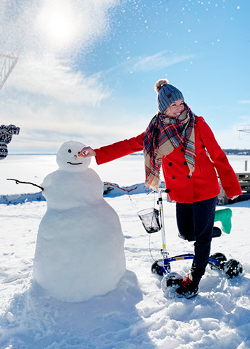 lady outdoor in the snow posing next to a snowman, she stands with her knee scooter