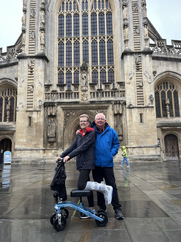 A smiling couple stands in front of Bath Abbey, with one person using a knee scooter, demonstrating mobility in front of the historic cathedral.
