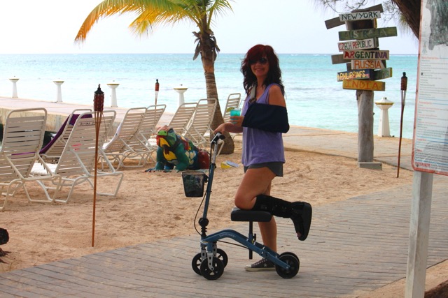 A woman with red hair, wearing a blue top and purple shorts, stands with a knee scooter on a sandy beach area with palm trees and directional signs in the background.