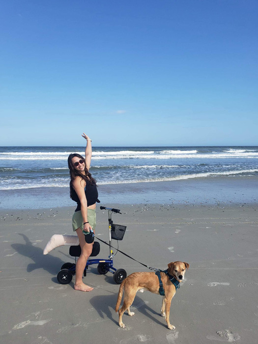 female patient riding a knee scooter while walking her dog, they take a stroll on the beach shore