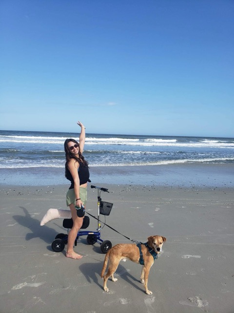 A young woman with dark hair, wearing sunglasses, a tank top, and shorts, joyfully poses on a beach with a leashed dog and a mobility scooter.