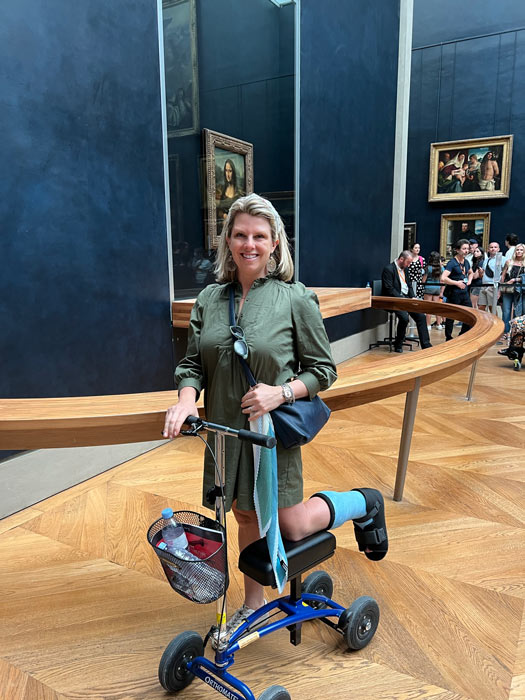 woman using a knee scooter inside the Louvre museum in Paris. Behind her is the Mona Lisa painting.