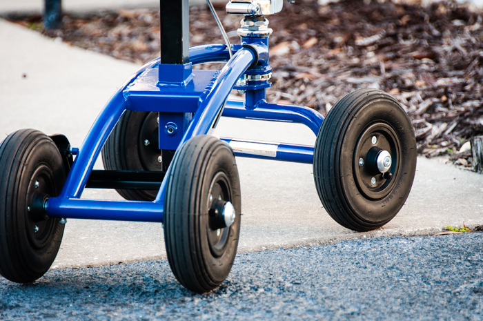 Close-up of a blue knee scooter showing its air-filled tires and sturdy metal frame, positioned on a concrete surface with mulch visible in the background.