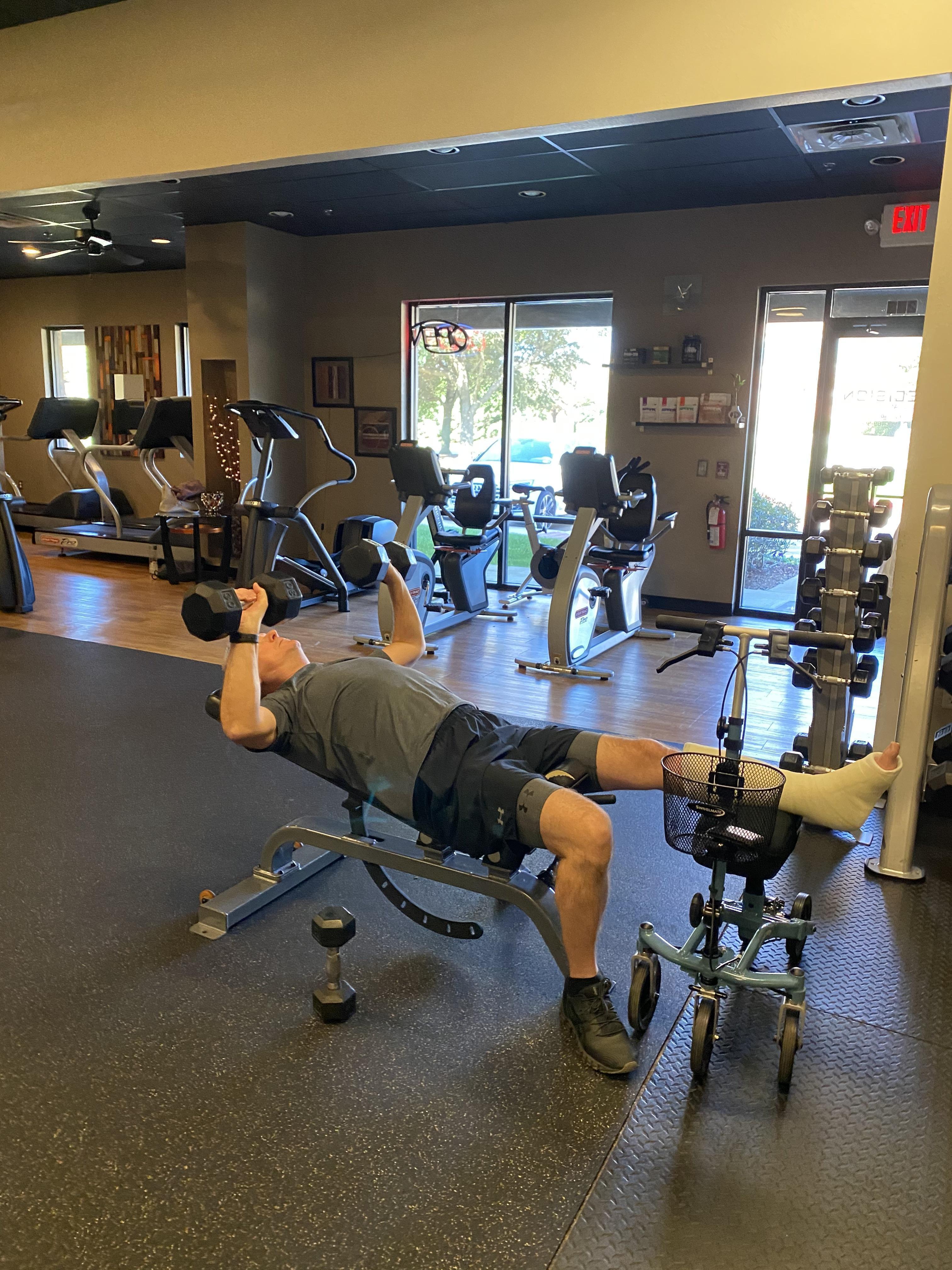 A man lifts light weights using his knee scooter for support, showcasing resilience and determination in maintaining an active lifestyle.