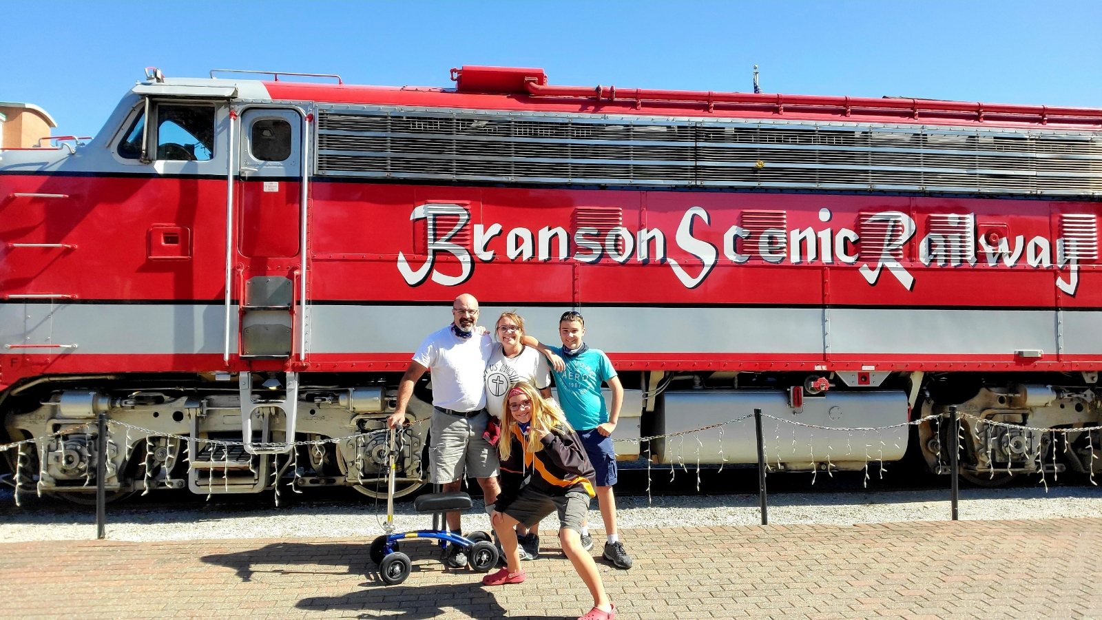 In front of a picturesque train, a father with a beaming smile poses proudly with his three children, all gathered around his knee scooter. The father, equipped with his mobility aid, exudes strength and love as he embraces his kids. Each child wears a joyful expression, radiating happiness and a sense of togetherness. The train, a symbol of adventure and journey, adds a touch of excitement to the background. This heartwarming scene captures the bond between a caring father and his playful, adoring children, as they create lasting memories together.