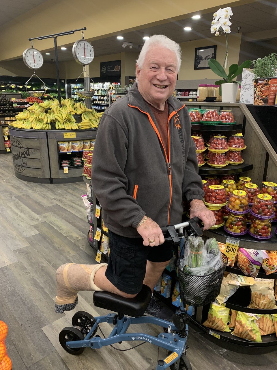 A senior with a leg injury navigates the aisles of a bustling supermarket on a knee scooter. He wears a determined expression as he carefully selects fresh produce from the shelves. His knee scooter, a mobility aid with handlebars and wheels, allows him to move around comfortably while keeping weight off his injured leg. Brightly colored fruits and vegetables surround him, offering a vibrant and nutritious array of options. The scene captures his independence and resilience in adapting to his circumstances during the shopping trip.