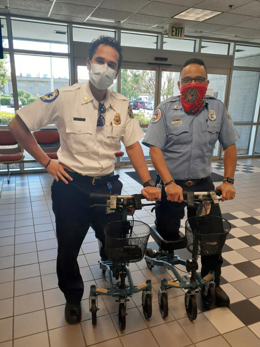 Two emergency medical dispatch (EMD) paramedics wearing uniforms and helmets, standing side by side. They each have a knee scooter, a mobility aid with wheels designed to support their injured legs or feet.