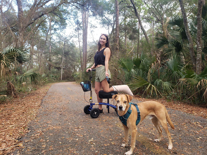  In a peaceful nature trail, a woman gracefully rides her knee scooter while her loyal dog walks alongside her.