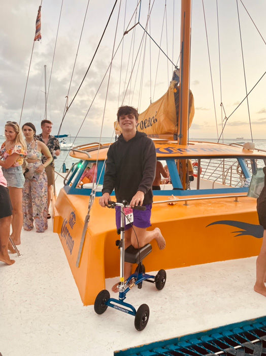 Enjoying a sailing adventure, a young boy stands on a sailboat, his Orthomate knee scooter by his side, as the wind fills the sails and the water glistens beneath them.