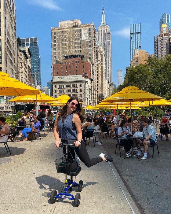  In the bustling streets of New York City, a young lady confidently navigates her knee scooter. The Empire State Building stands tall in the background, creating a picturesque urban backdrop.